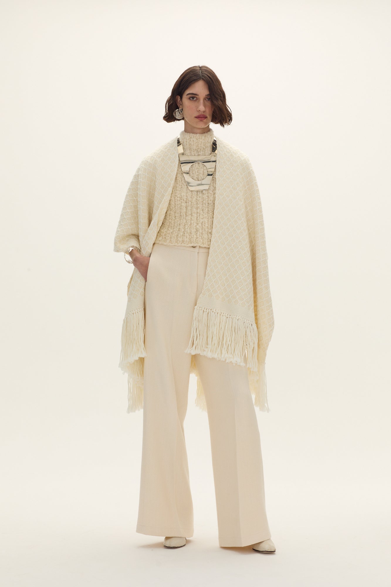 A model wearing a beige hand loomed ruana with tassels, paired with matching trousers and a sweater.