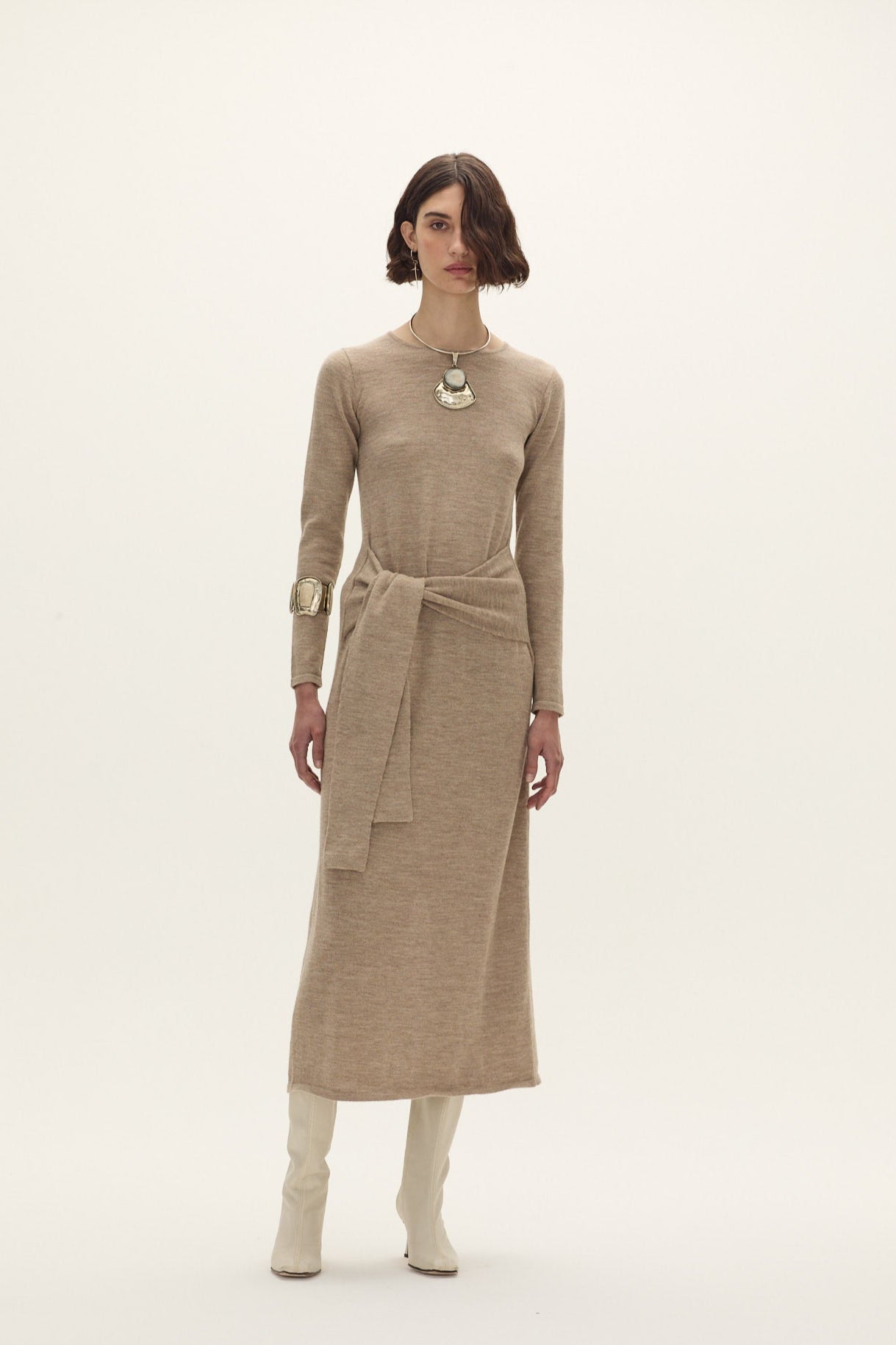 Woman wearing a A long, fitted dress with straight lines. Waist tie at the front. Long sleeves.